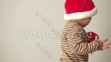 戴圣诞帽的可爱宝宝手里拿着一个红色的圣诞球。 准备圣诞晚会，快乐的诺埃尔